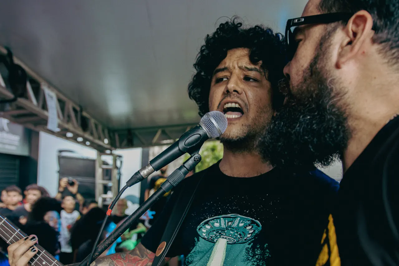 Dois músicos cantam no mesmo microfone, rosto a rosto.